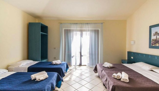 Camera spaziosa con tre letti, copriletti blu e marroni, e tendaggi chiari.