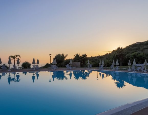 Piscina tranquilla al tramonto, con ombrelloni e palme visibili sullo sfondo.