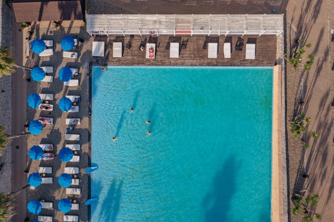 Veduta aerea di una piscina con ombrelloni blu e lettini bianchi. Alcune persone nuotano.