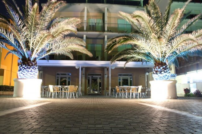 Ingresso di un hotel illuminato di sera, con palme e tavolini.