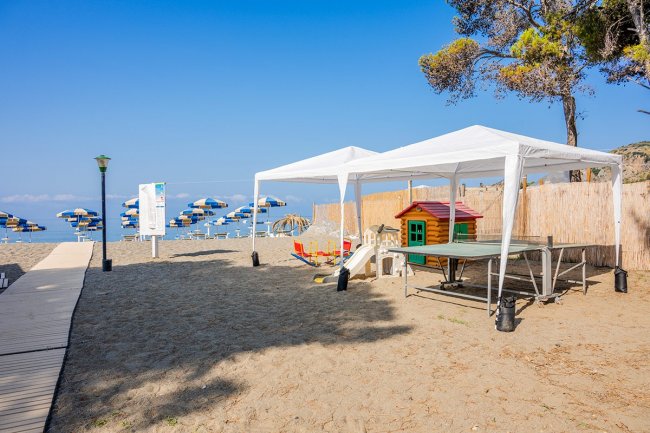 Area gioco sulla spiaggia con tenda bianca e casetta.