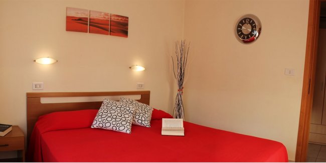 Letto matrimoniale con copriletto rosso e cuscini decorativi.