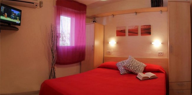Camera da letto con letto rosso, cuscini decorativi e televisore.