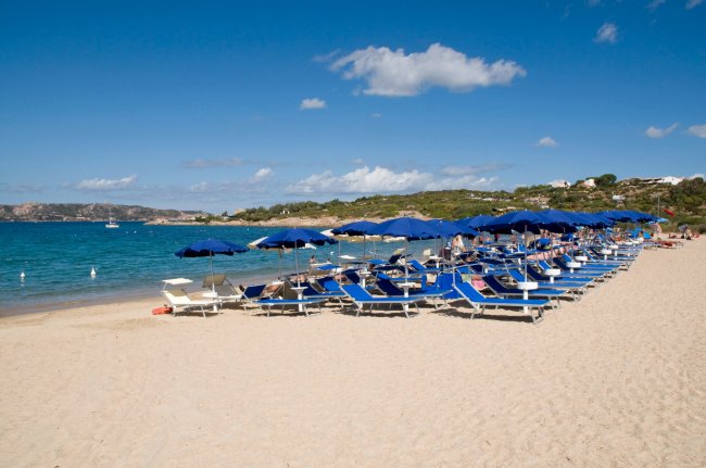 Spiaggia con sabbia chiara, ombrelloni blu e lettini, mare calmo e cielo azzurro.