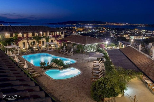 Vista notturna di un hotel con due piscine illuminate e città in lontananza.