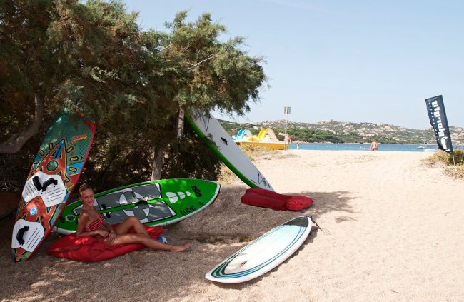 Una donna seduta su un cuscino rosso sotto un albero vicino a tavole da surf sulla sabbia.