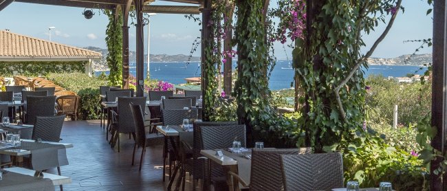 Veranda di un ristorante con sedie e tavoli, vista mare e vegetazione rampicante.